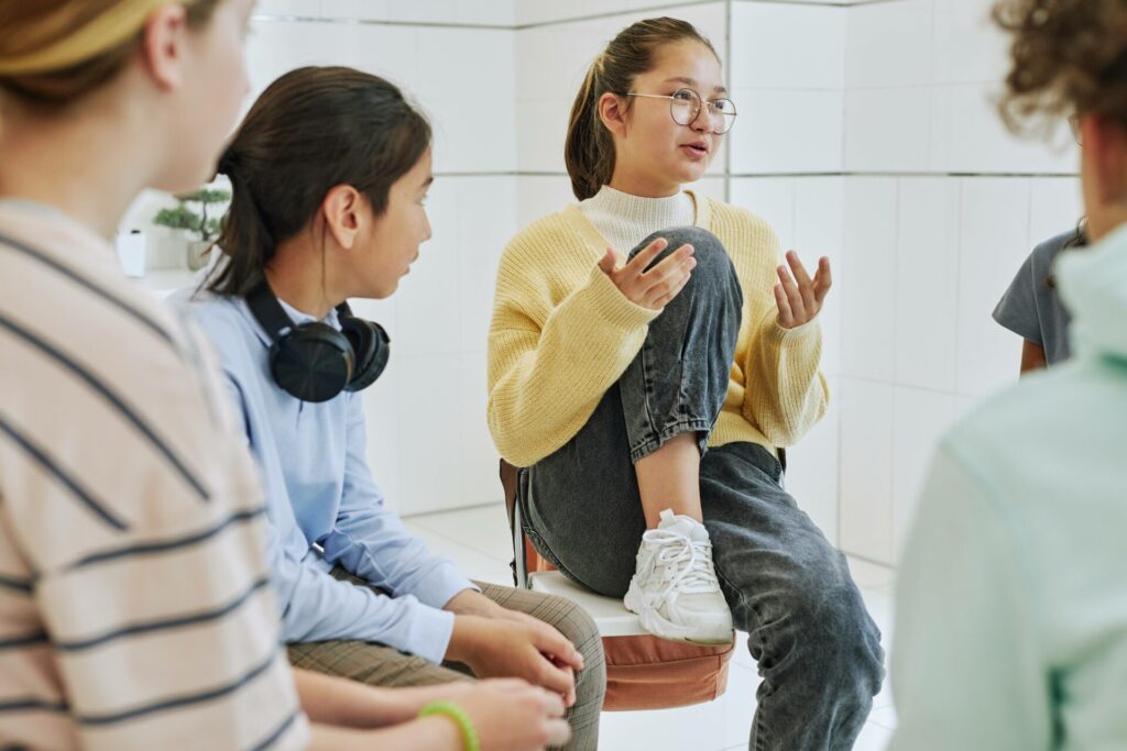 Groupe d’adolescents en discussion dans un cadre scolaire ou éducatif. Une jeune fille, assise sur une chaise, prend la parole avec expressivité pendant que les autres écoutent attentivement. L’ambiance est détendue et propice à l’échange, illustrant un atelier de parole, un cercle de discussion ou une activité pédagogique participative.