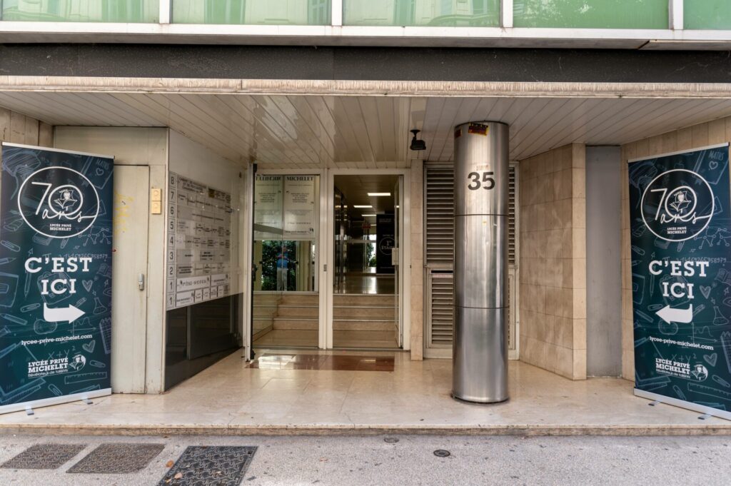 Entrée du lycée privé Michelet à Nice, située dans un immeuble urbain. Deux kakémonos signalent les 70 ans de l’établissement avec la mention "C’est ici". L'entrée est propre et accueillante, avec une colonne métallique portant le numéro de rue. On aperçoit l’intérieur du bâtiment à travers les portes vitrées.