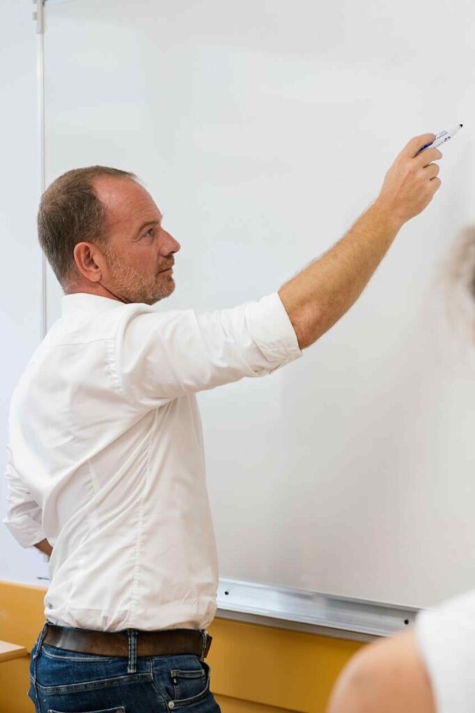Dans une salle de classe, un professeur en chemise blanche se tient debout devant un tableau blanc et lève le bras avec un feutre pour écrire ou expliquer un point du cours.