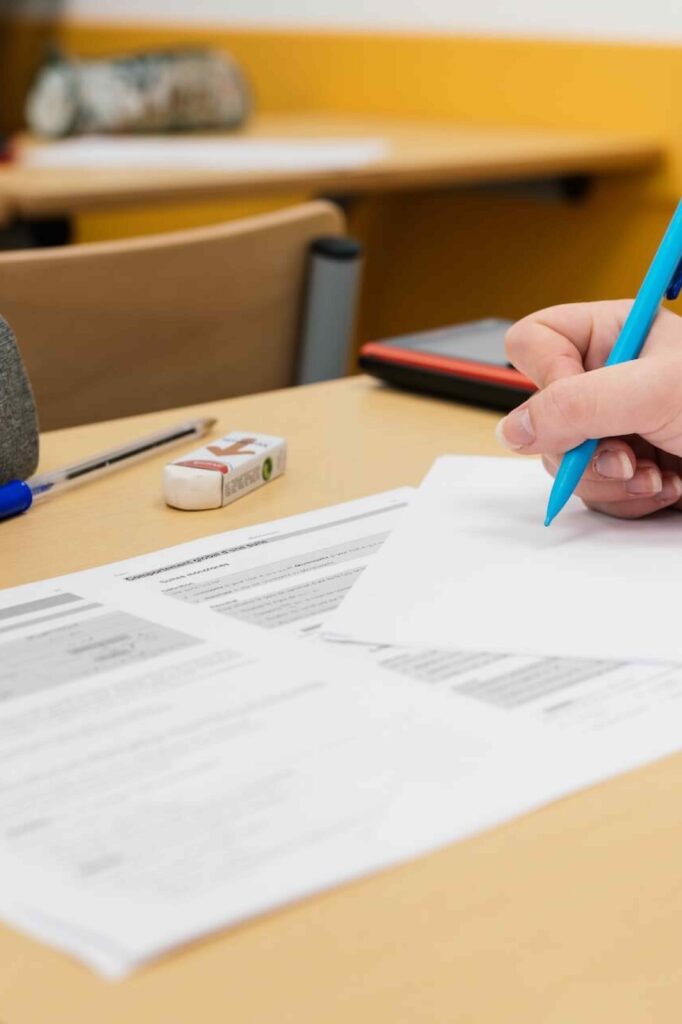 Vue rapprochée d’une main écrivant sur une feuille blanche posée sur un bureau de classe, à côté de documents imprimés, d’une gomme, d’un stylo et d’une calculatrice.