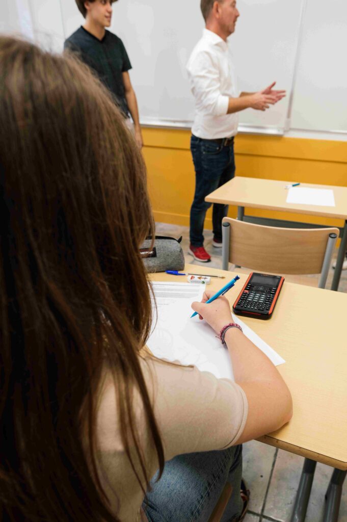 Vue arrière d’une élève assise en salle de classe, écrivant sur une feuille à son bureau, tandis qu’un enseignant et un autre étudiant se tiennent devant le tableau blanc.