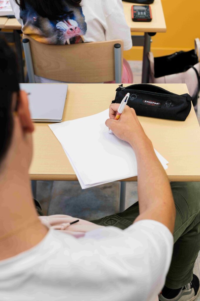 Vue rapprochée d’un élève assis en salle de classe, écrivant sur une feuille blanche à son bureau, avec une trousse noire et un ordinateur portable à proximité.